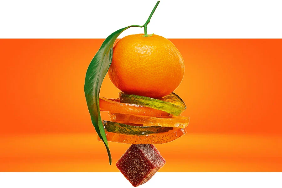 Photographie d'une mise en scène avec une orange empilée sur des fruits confits et un cube de pâte de fruit 'Dés Lisses' de la marque Cruzilles